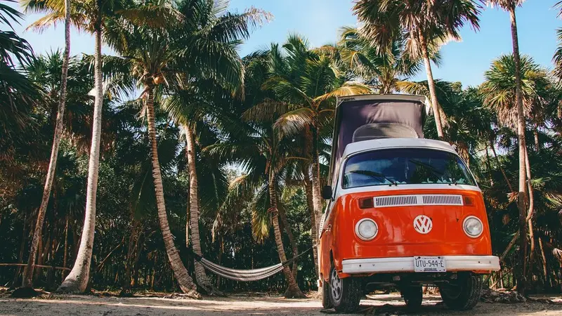 Klassischer orangefarbener VW-Kombi mit geöffnetem Aufstelldach zwischen Palmen neben einer Hängematte